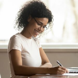 A woman ordering home services online at clickapro.com and saving time and money. The woman is sitting at a kitchen table and is holding a pen and writing on a piece of paper. She is happy. She is wearing glasses and her dark curly hair is tied back.