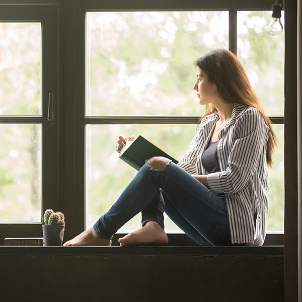 A woman sitting near her window reading a book. The light coming through the window is soft because the window is protected by a solar window screen.