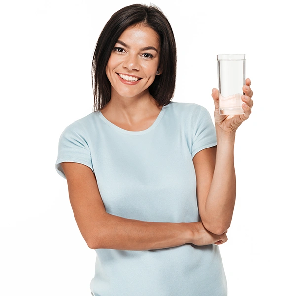 A woman in a light blue shirt and dark hair holding up a glass of water. She is calculating her household's water use to plan for her hauled water needs